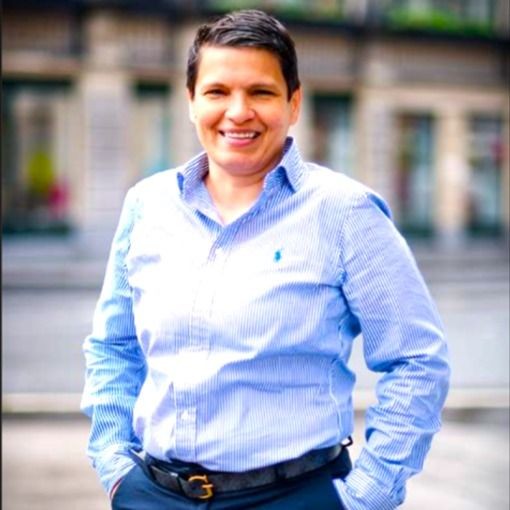 Person smiling and standing outdoors, wearing a light blue button-down shirt and dark pants.